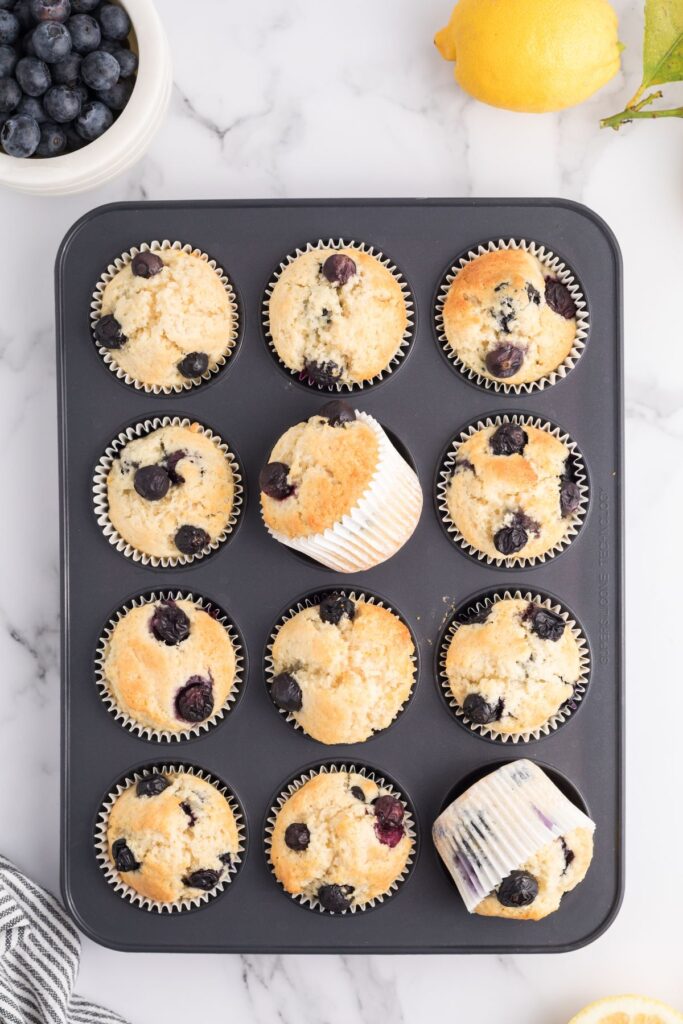 Golden muffins in the muffin tin after being baked, with blueberries and lemons on the table. 