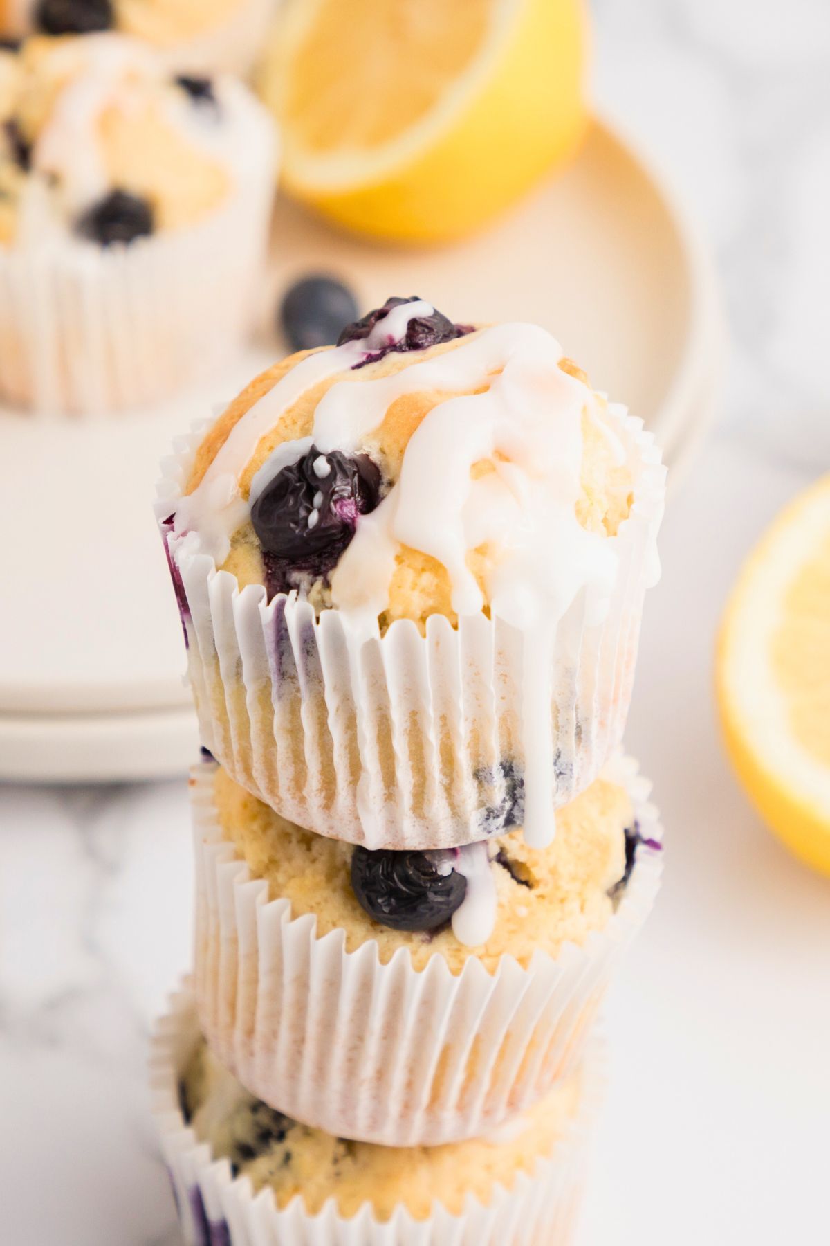 Lemon blueberry muffins with white icing drizzle, stacked on a white marble table.