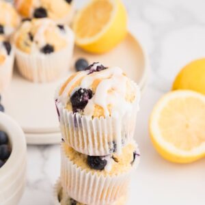 Golden lemon blueberry muffins baked and then stacked on the table with lemons stacked behind the muffins.