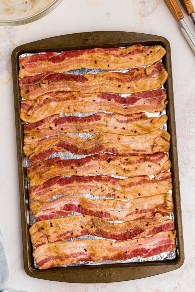 Uncooked bacon that has been coated with sugar and spices on a baking sheet lined with foil.