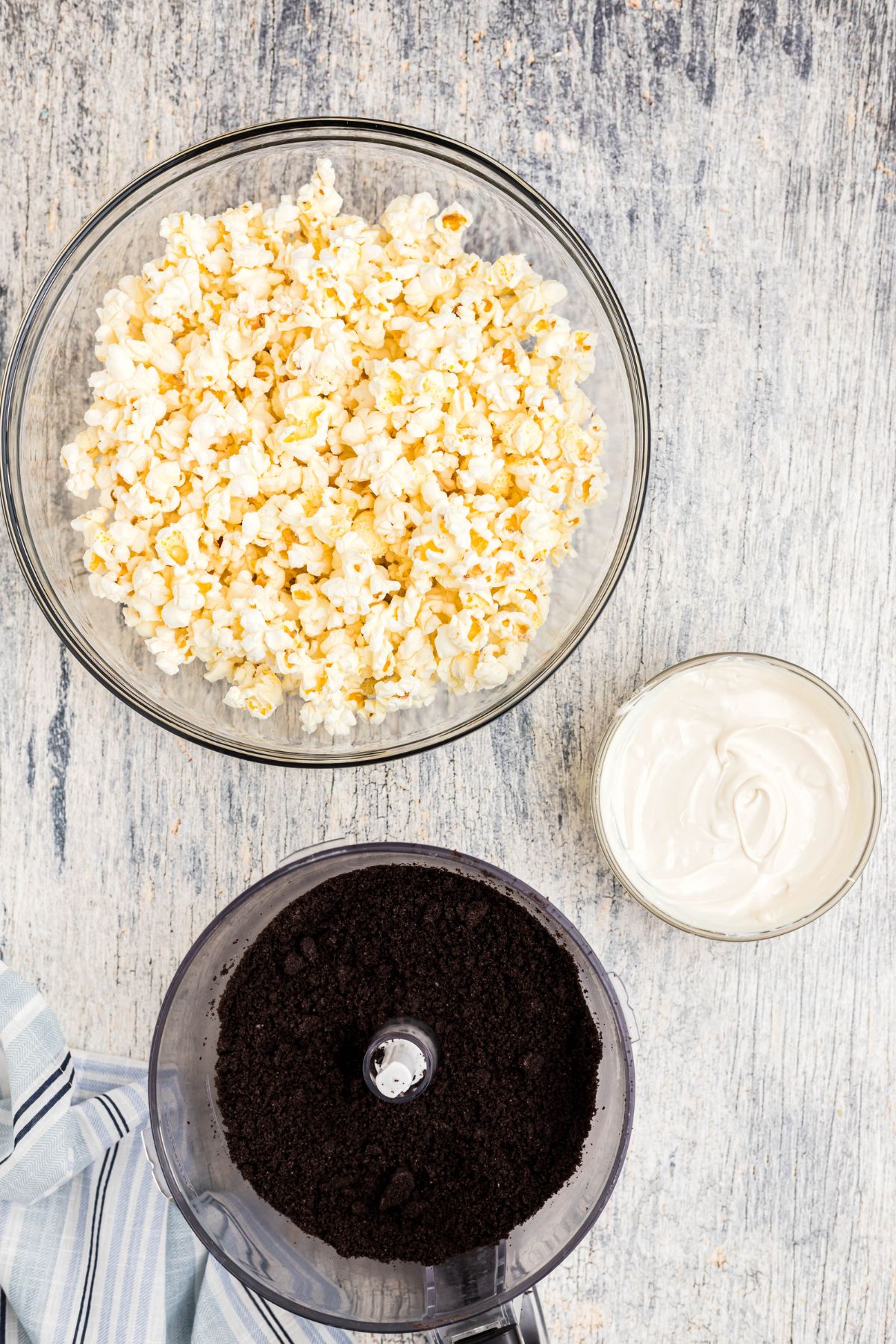 Oreos in a food processor bowl, crushed into crumbs, with melted white chocolate and a bowl a popcorn, on a blue wooden table. 