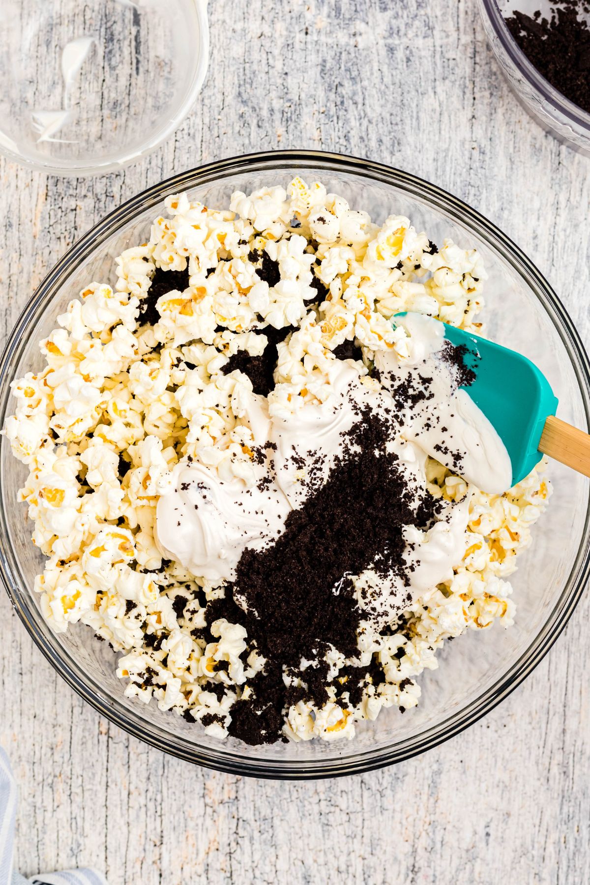 Melted white chocolate with Oreo crumbles being mixed in a clear glass bowl filled with popcorn, with a spatula. 