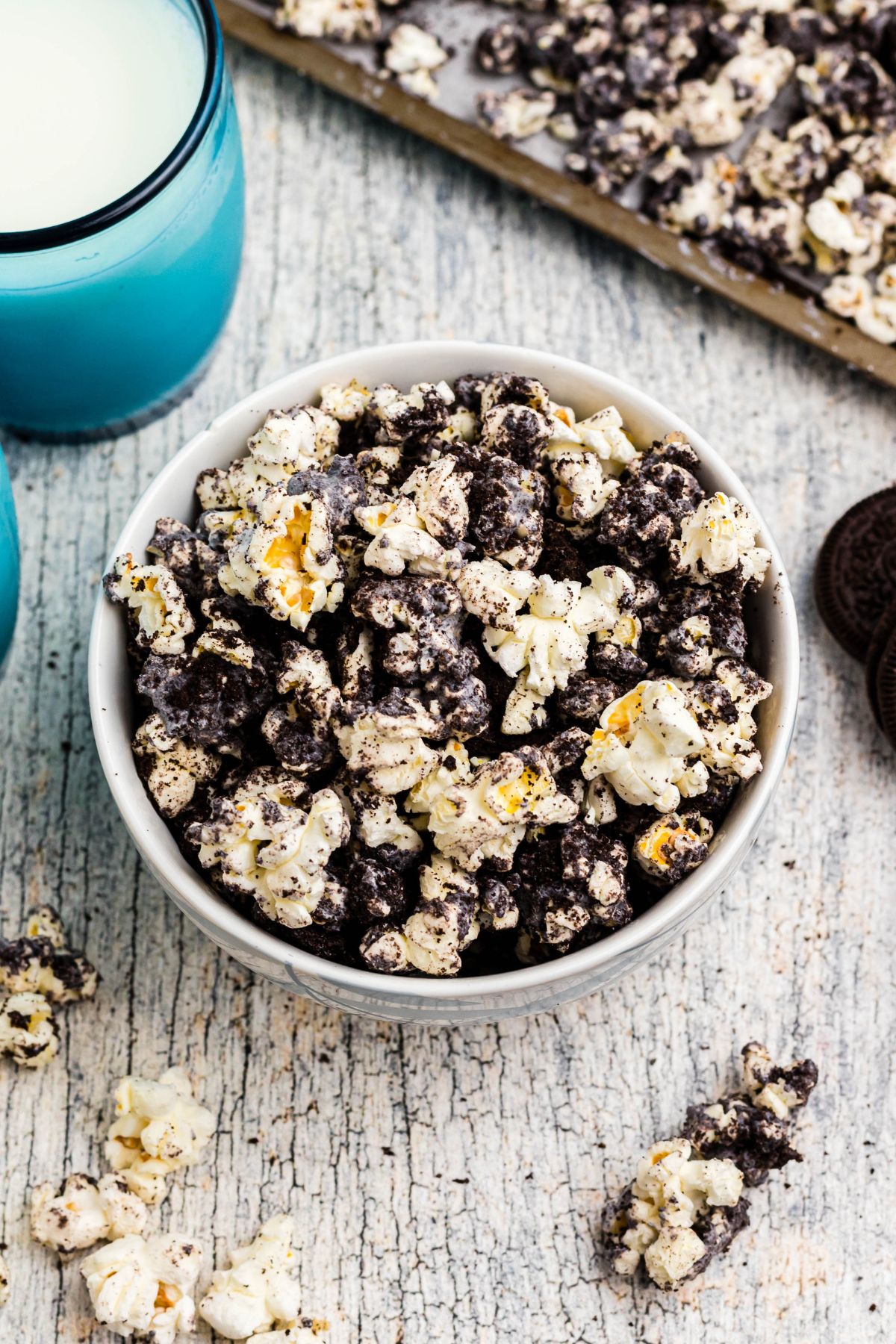 A bowl filled with Oreo popcorn on a table with blue glasses filled with milk and popcorn pieces scattered on the table. 