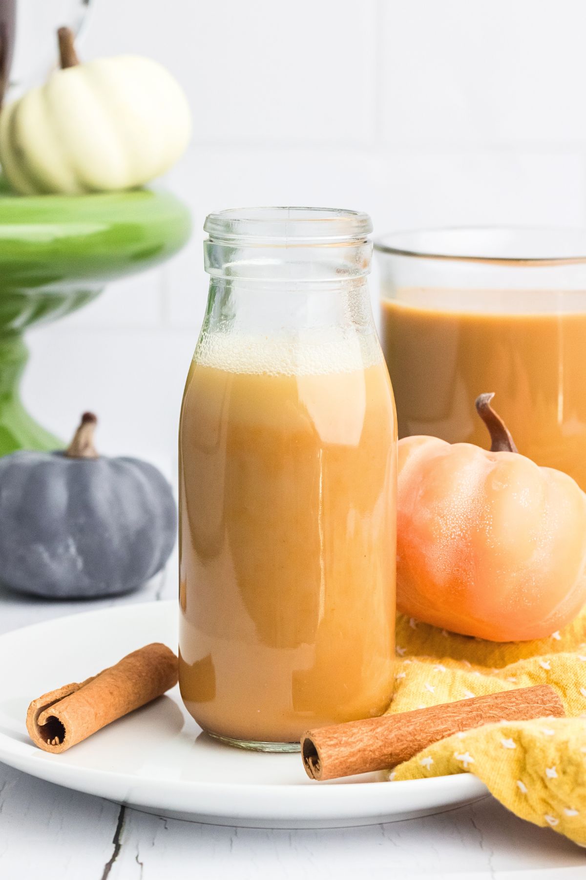 Pumpkin spice creamer in a small bottle on a white plate with cinnamon sticks and small pumpkins on the table. 