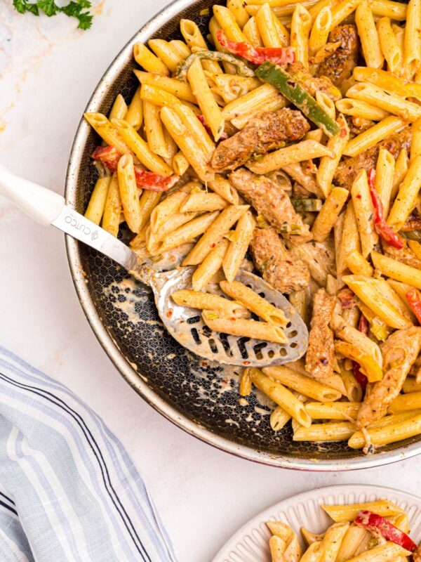 Cooked pasta topped with cheesy cream sauce and saut&eacute;ed veggies and seasoned chicken in a large skillet with a large serving spoon in the center.