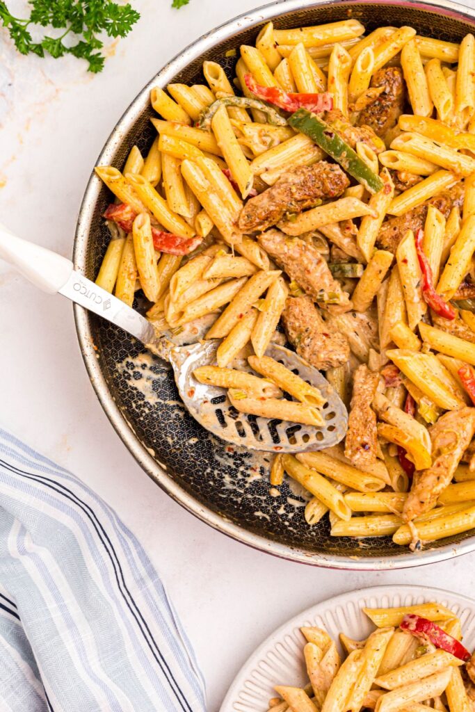 Serving spoon in the middle of skillet of pasta and cooked chicken and vegetables. 