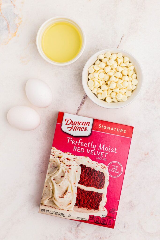 Box of red velvet cake mix with white chips, eggs, and oil on a marble table. 