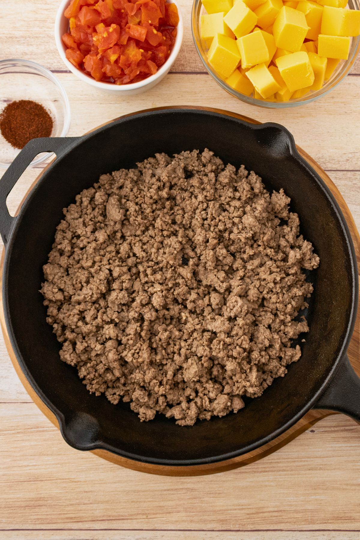 Ground beef cooked in a black skillet with the grease drained and cheese cubes, chopped tomatoes, and seasonings on the table. 