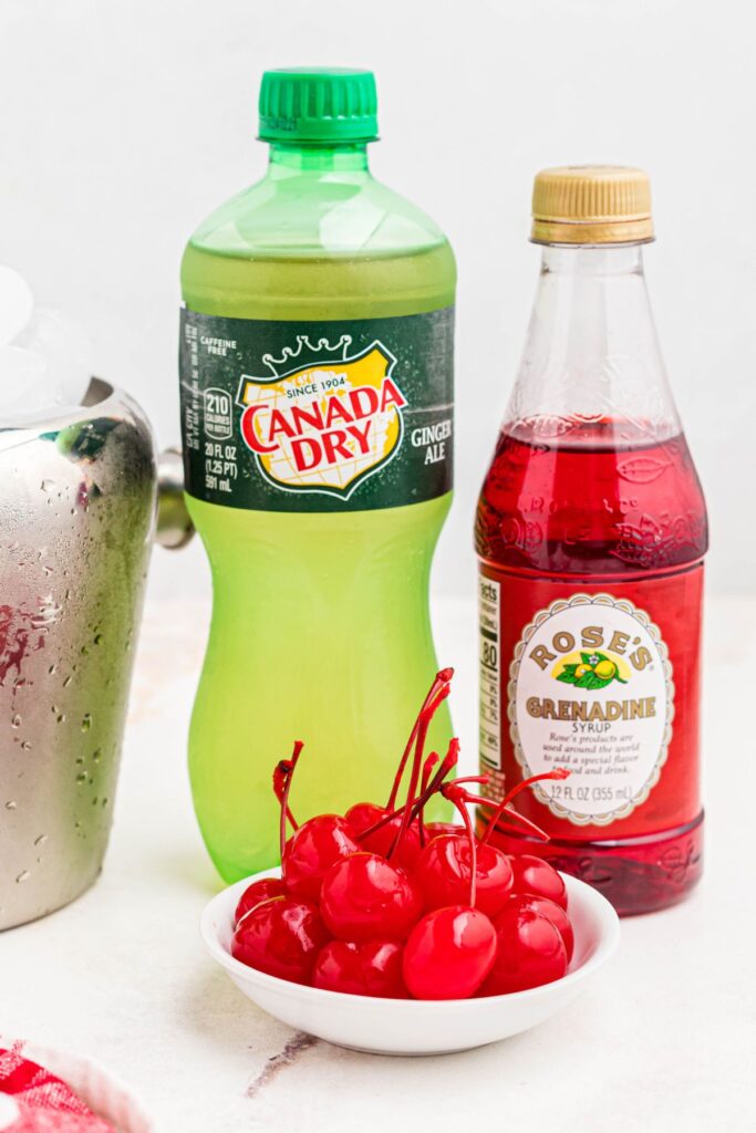 Ingredients needed to make Shirley temple drink of Ginger ale, grenadine, and cherries on a marble table. 