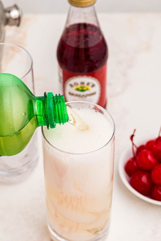 Ginger ale being poured over ice on a marble table, with a small bowl of cherries next to the glass. 