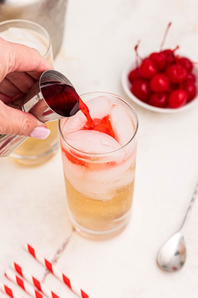 A Shirley Temple drink being mixed with ingredients of red grenadine being poured into a tall glass over ice. 