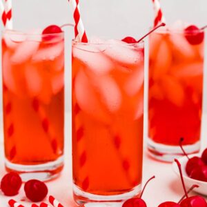 Bright red drink in a tall glass with ice and cherries on the table.