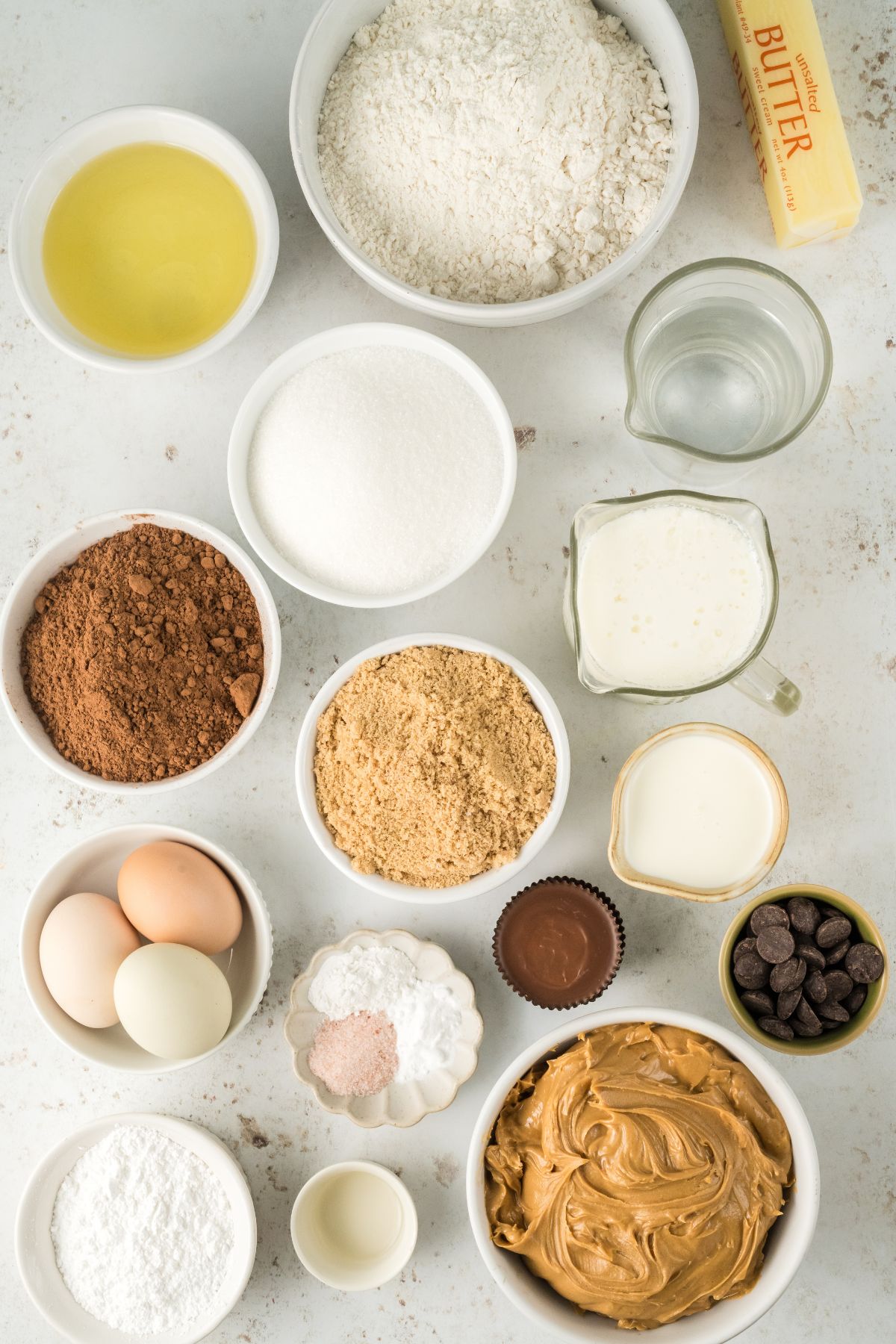 Ingredients needed to make chocolate peanut butter cake measured out in small glass bowls on a marble table. 