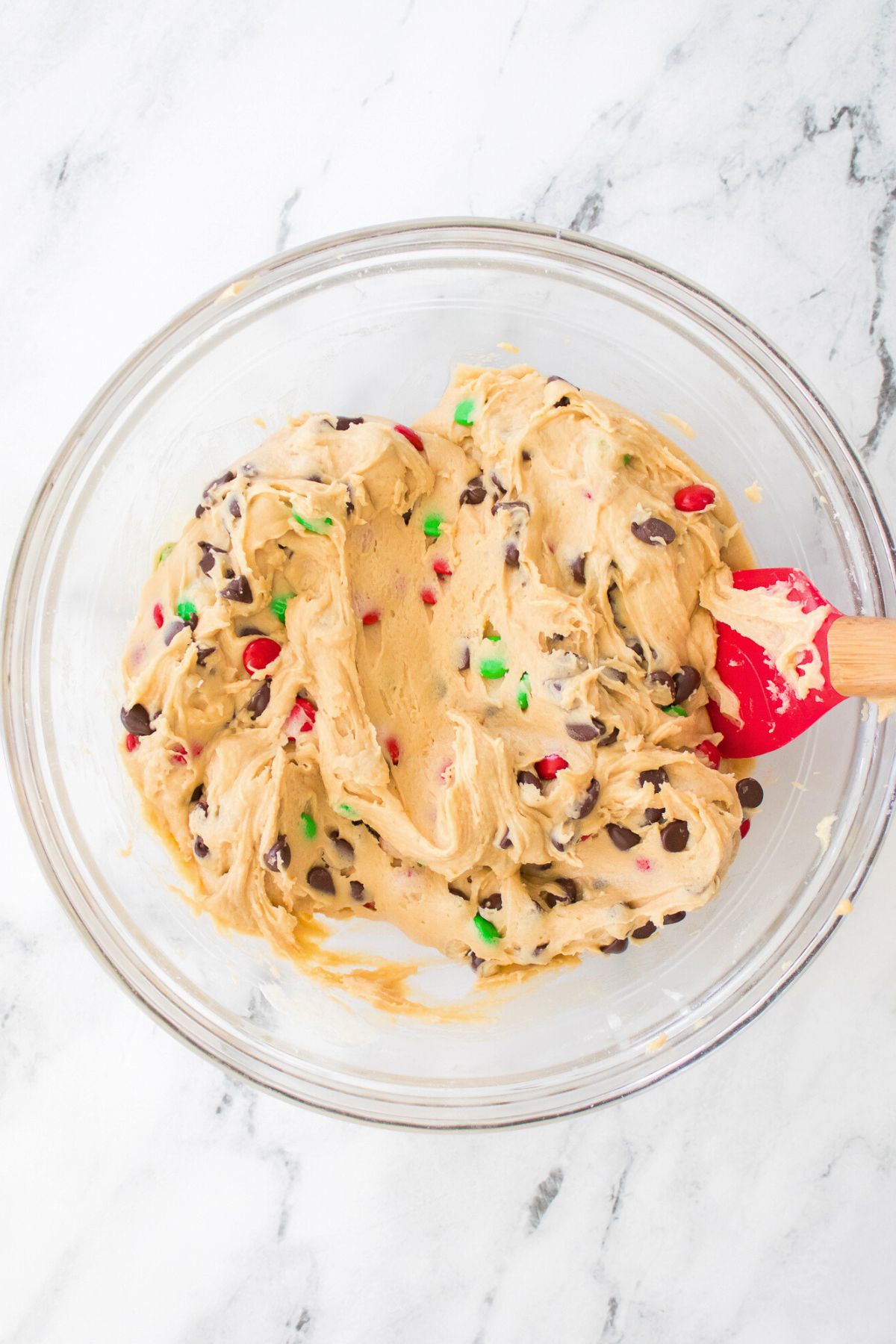 Sugar cookie dough with candies and chocolate chips mixed in, inside of a large clear glass bowl. 