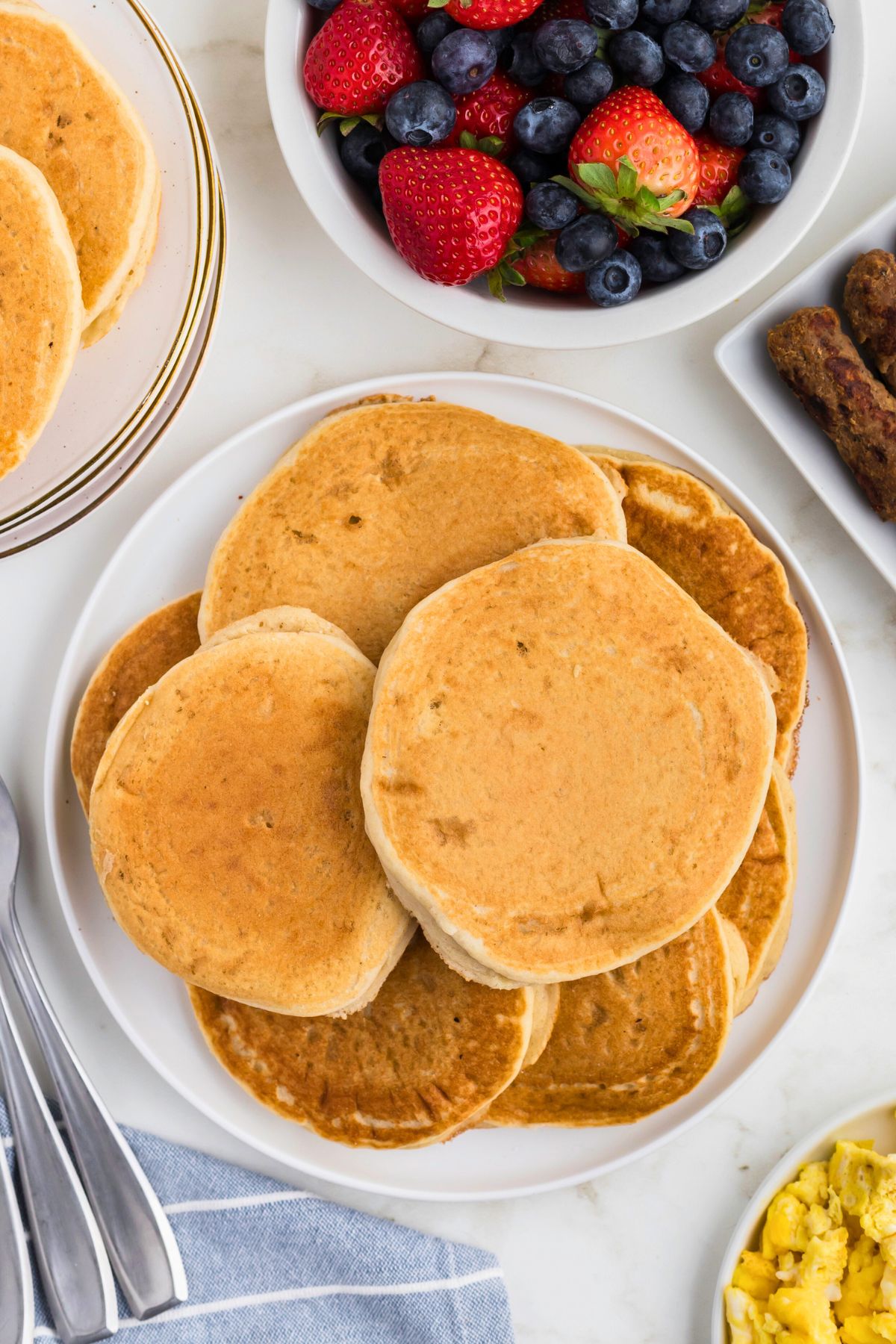 Light and fluffy pancakes stacked on a plate with fruit and sausage on the table. 