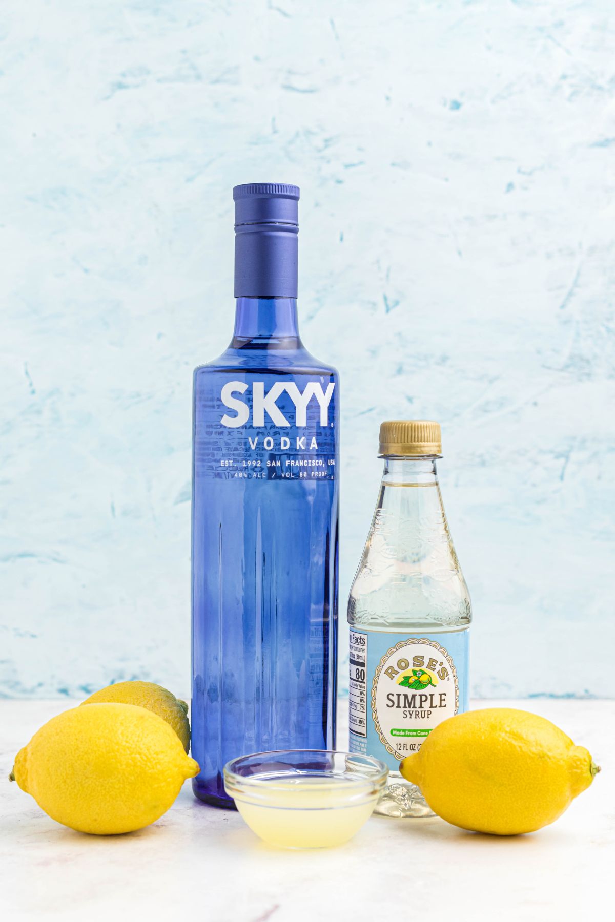 Bottles of vodka and simple syrup, with a small bowl of lemon juice on a marble table. 