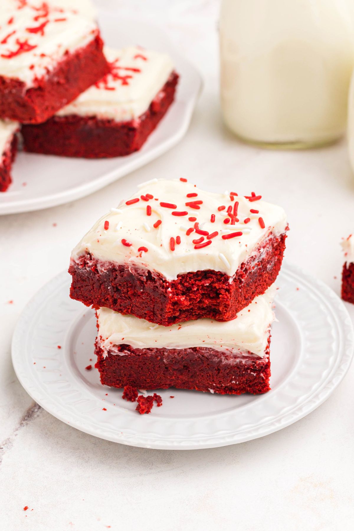 Red velvet bars frosted with cream cheese frosting, stacked on a white plate, one with a bite missing. 