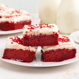 Red velvet bars frosted with cream cheese frosting, stacked on a white plate.