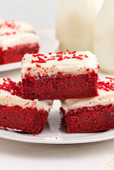 Red velvet bars frosted with cream cheese frosting, stacked on a white plate.