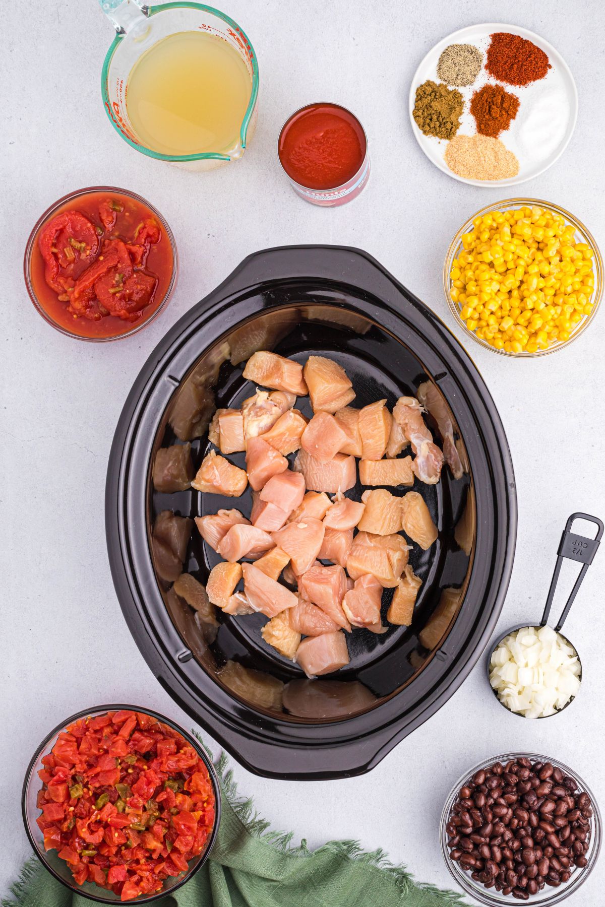 Chicken pieces in the bottom of a black crockpot bowl with other chili ingredients on the table. 