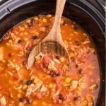 Chicken chili in a black slow cooker with a wooden spoon serving it.