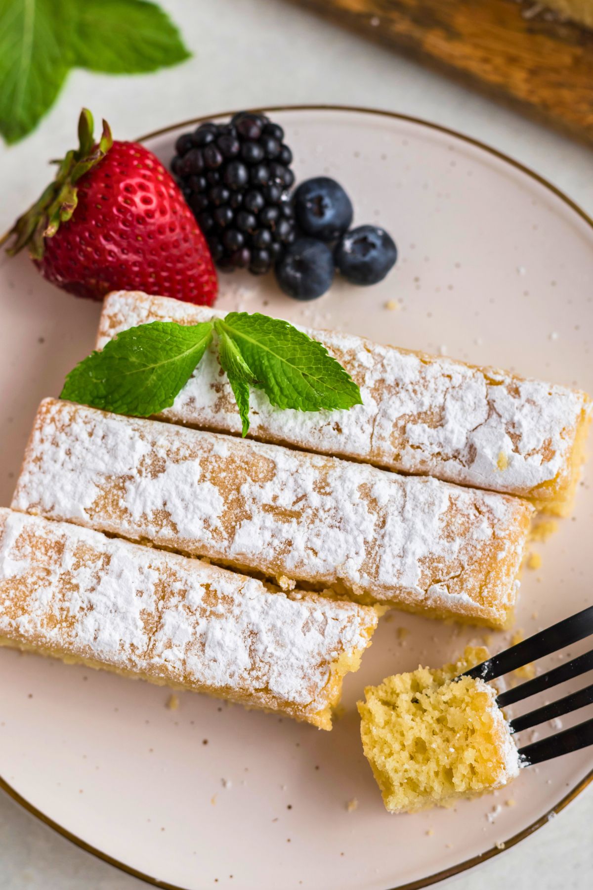 Golden yellow butter cake slices on white plates topped with powdered sugar with berries on the table, with a fork taking a small bite. 