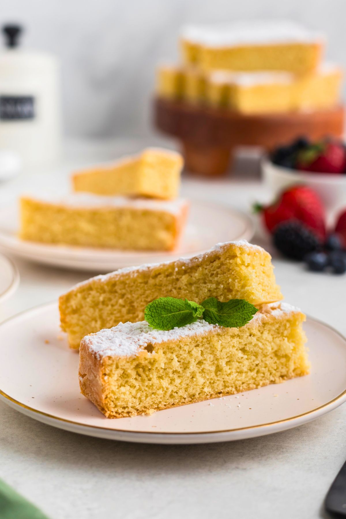 Golden yellow butter cake slices on white plates topped with powdered sugar with berries on the table. 