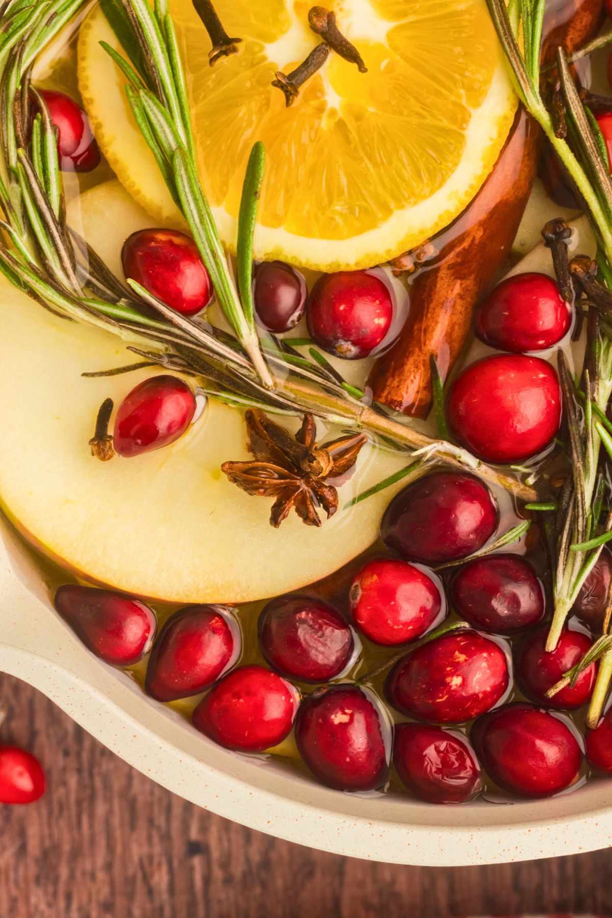 Cream colored saucepan filled with water, cranberries, fruit slices, and other ingredients to make a Christmas simmer pot.