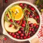 Cream colored saucepan filled with water, cranberries, fruit slices, and other ingredients to make a Christmas simmer pot on a wooden table with a red and white striped linen.