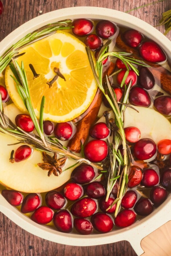 Cream colored saucepan filled with water, cranberries, fruit slices, and other ingredients to make a Christmas simmer pot on a wooden table with a red and white striped linen.