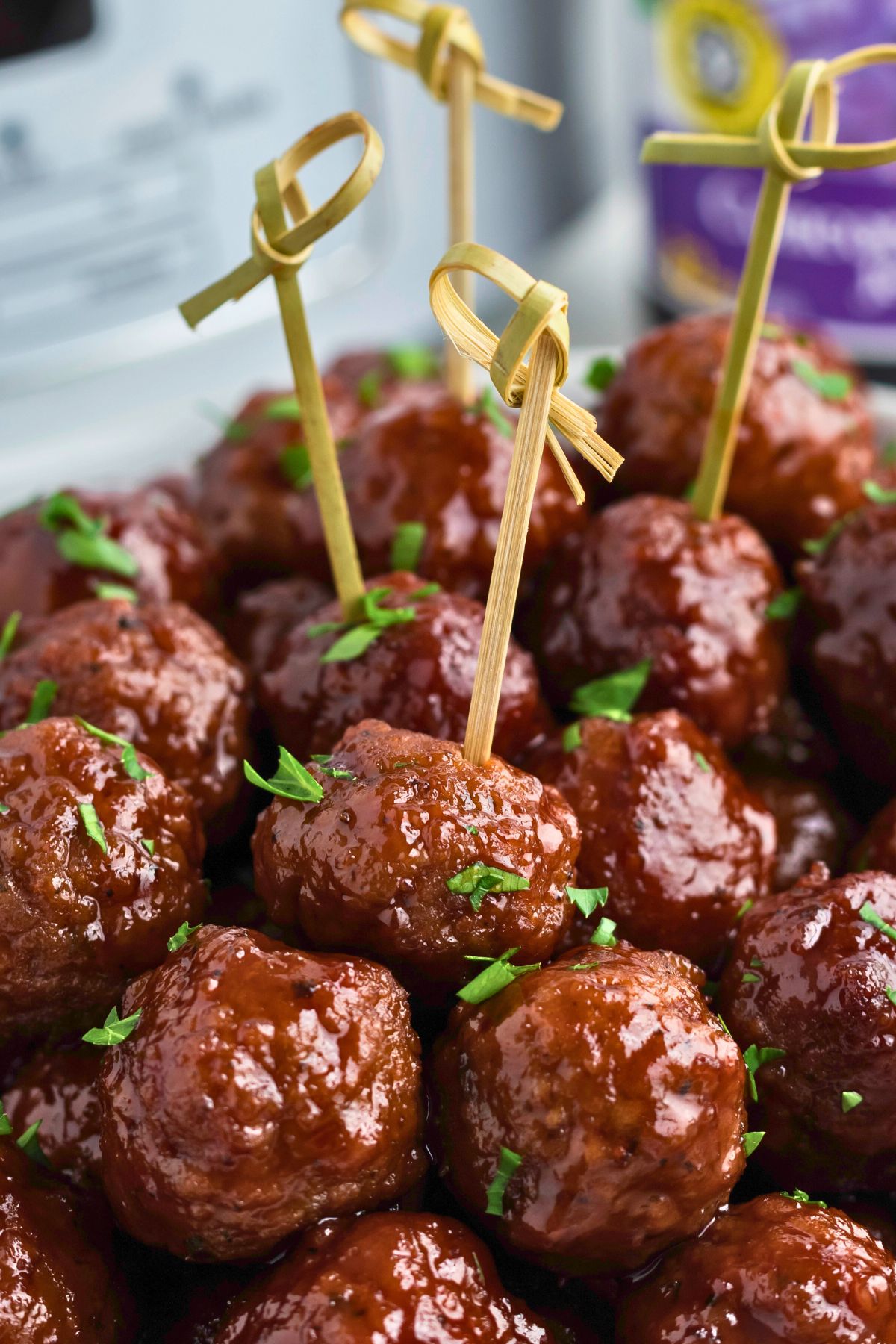 Grape jelly meatballs stacked with some skewered, in front of a slow cooker and jar of grape jelly. 