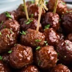 Grape jelly meatballs stacked with some skewered, in front of a slow cooker and jar of grape jelly.