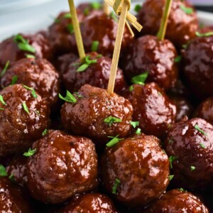 Grape jelly meatballs stacked with some skewered, in front of a slow cooker and jar of grape jelly.