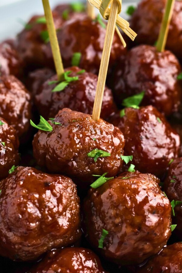 Grape jelly meatballs stacked with some skewered, in front of a slow cooker and jar of grape jelly.