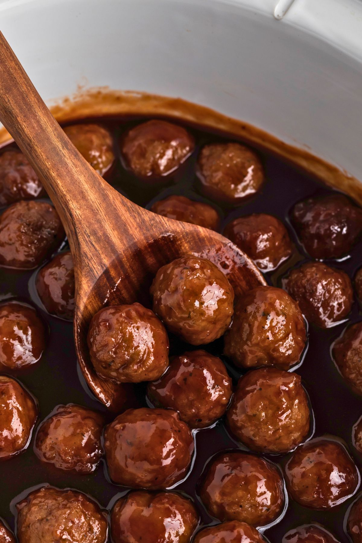 Grape jelly meatballs in a white crock pot being scooped up to serve with a wooden spoon. 