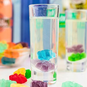 Purple and blue gummy bear in a clear shot glass with a vodka mixture poured over them, and with more gummy bears scattered on the table.