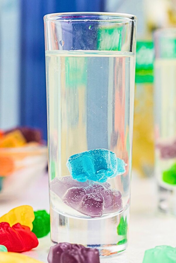 Purple and blue gummy bear in a clear shot glass with a vodka mixture poured over them, and with more gummy bears scattered on the table.