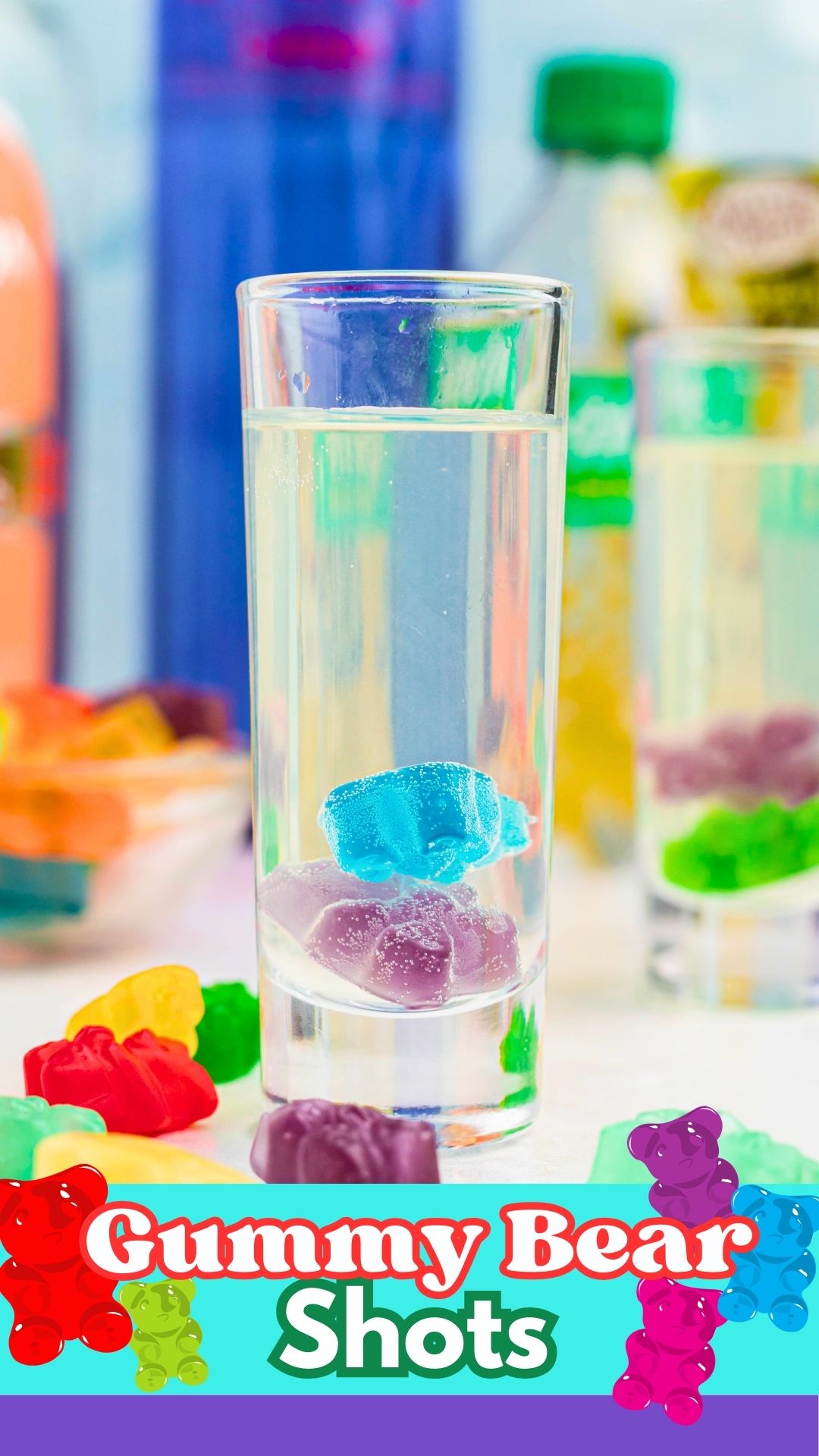 Purple and blue gummy bear in a clear shot glass with a vodka mixture poured over them, and with more gummy bears scattered on the table.