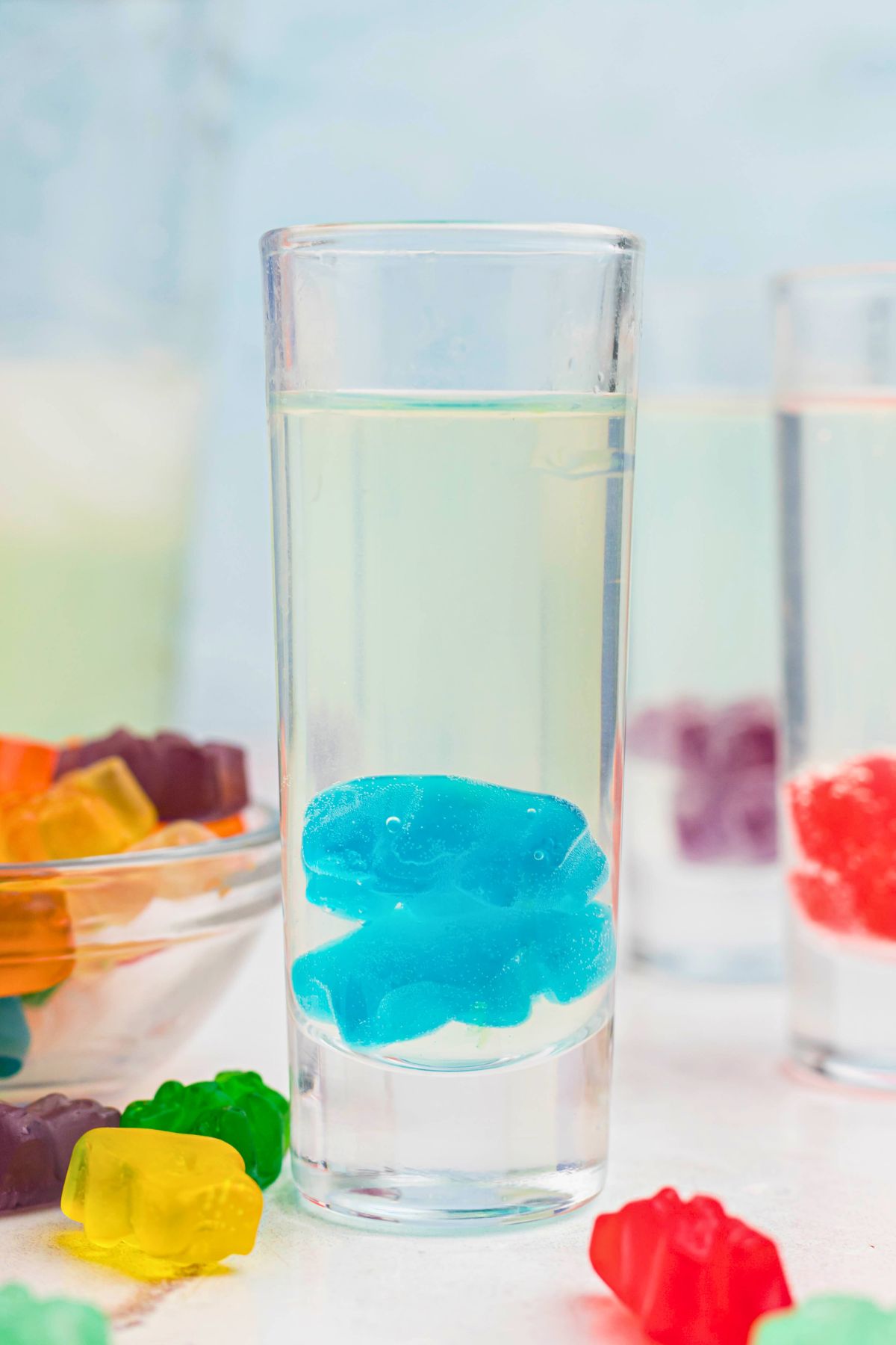 Shot glass filled with vodka and other liquids, with gummy bears dropped inside of the glass and on the table. 