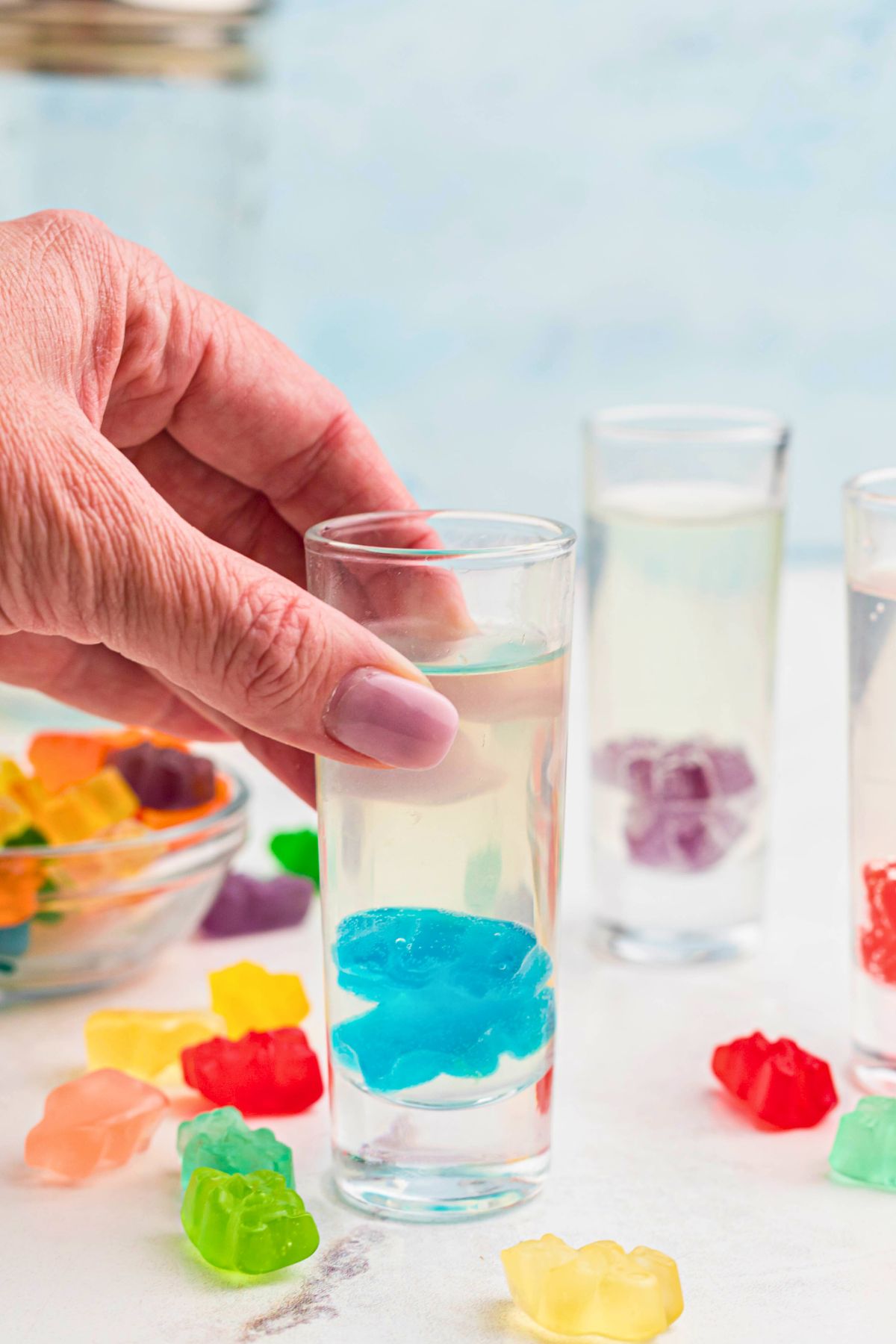 Blue gummy bears in the bottom of a shot glass with a hand picking up the glass. 