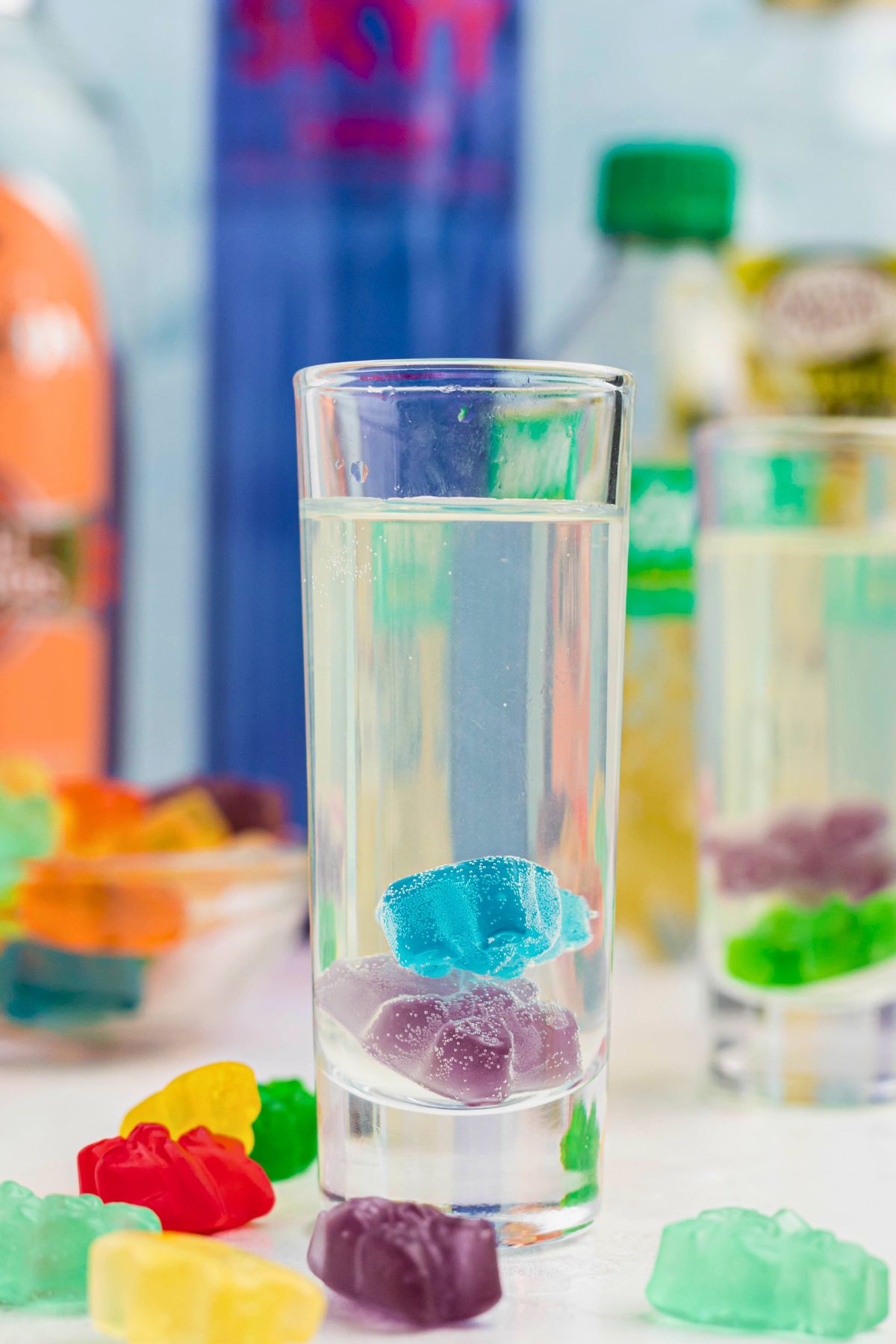 Purple and blue gummy bear in a clear shot glass with a vodka mixture poured over them, and with more gummy bears scattered on the table. 