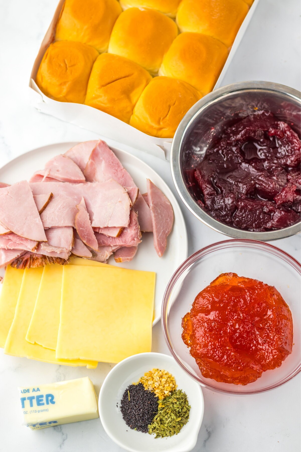 Ingredients needed to make ham and cranberry holiday sliders measured in bowls on a marble table. 