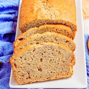 Golden loaf of banana bread on a white rectangle plate, sliced with a banana laying next to the plate.