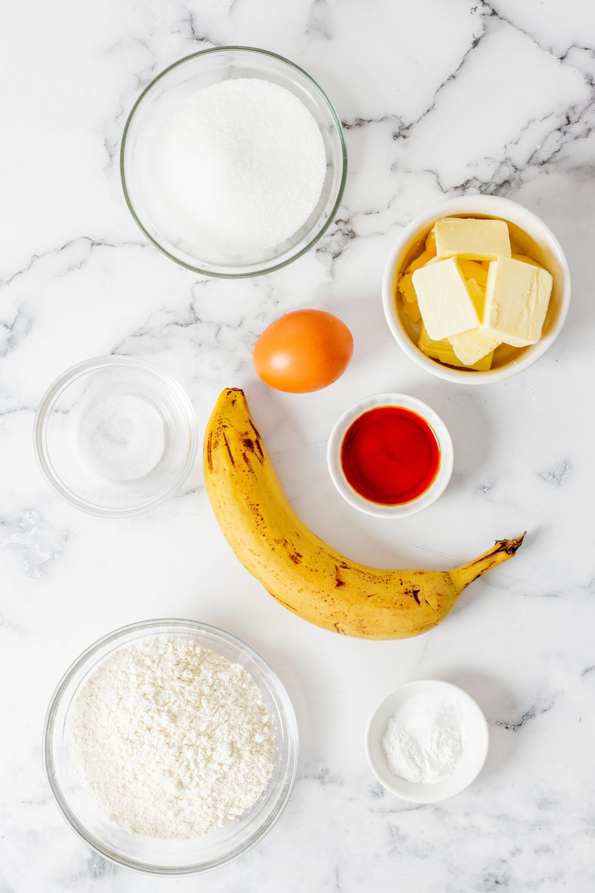 Ingredients needed to make banana bread measured into small glass bowls on a marble table. 