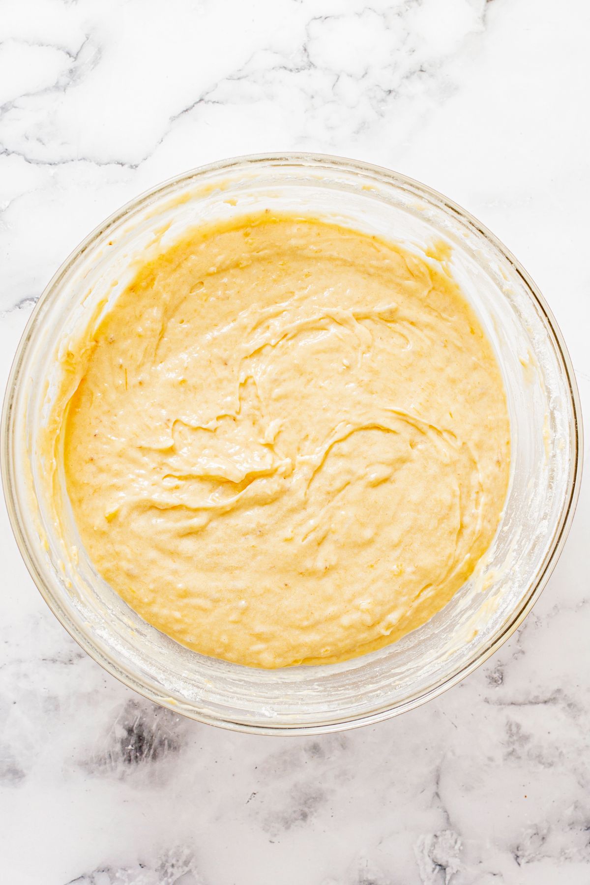 Banana bread batter in a large clear bowl on a marble table. 