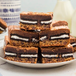 Stacked cookie brownies on a white plate with bottles of milk in the background on the table.