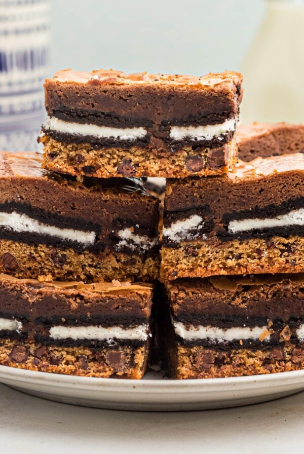 Stacked cookie brownies on a white plate with bottles of milk in the background on the table.