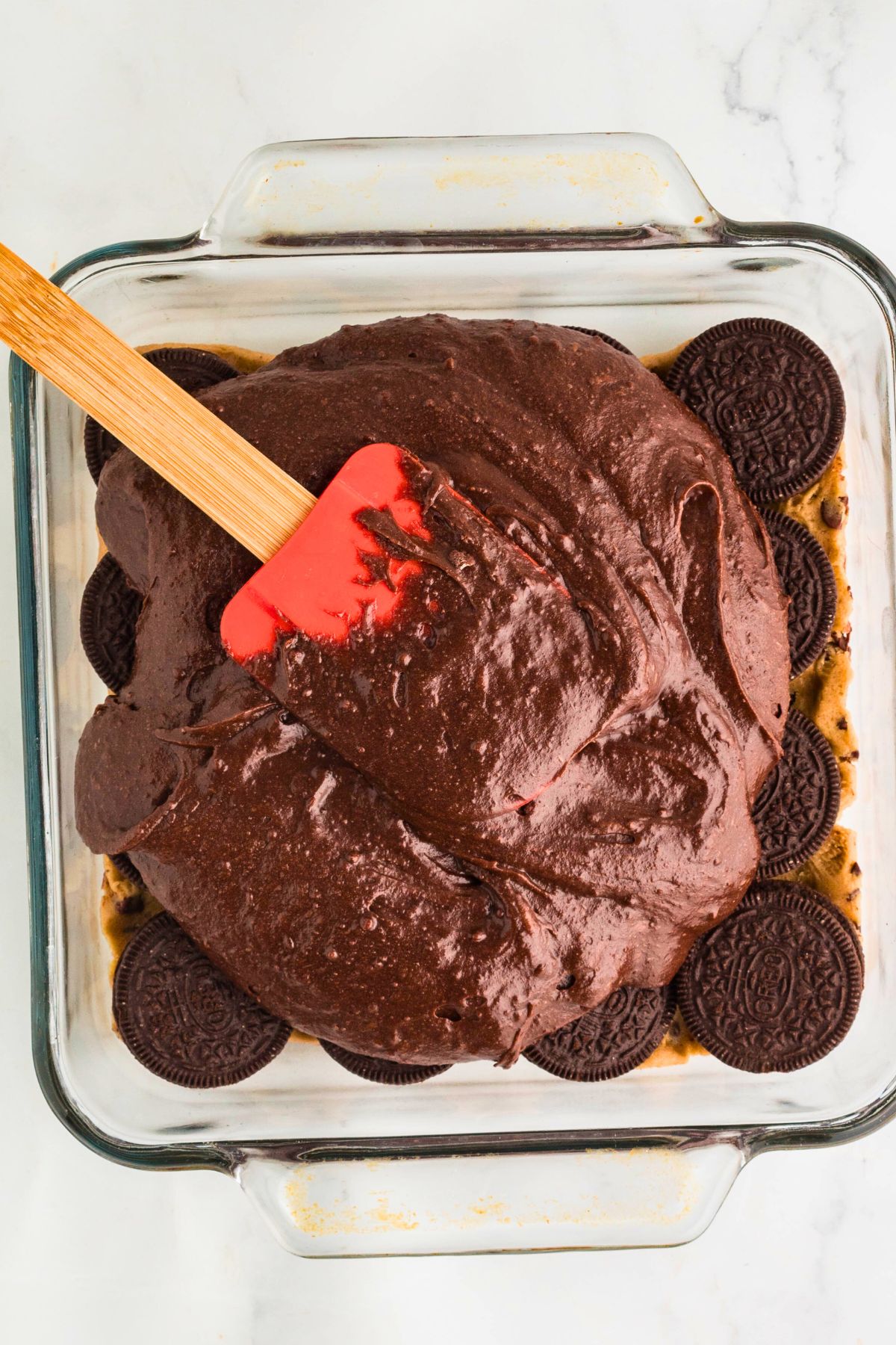 Brownie batter being spread over Oreo cookies and chocolate chip cookie dough in a baking dish. 