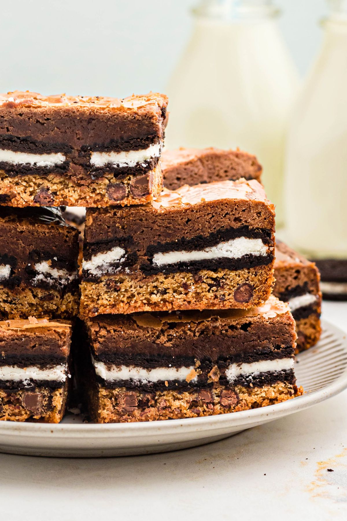 Stacked cookie brownies on a white plate with bottles of milk in the background on the table. 