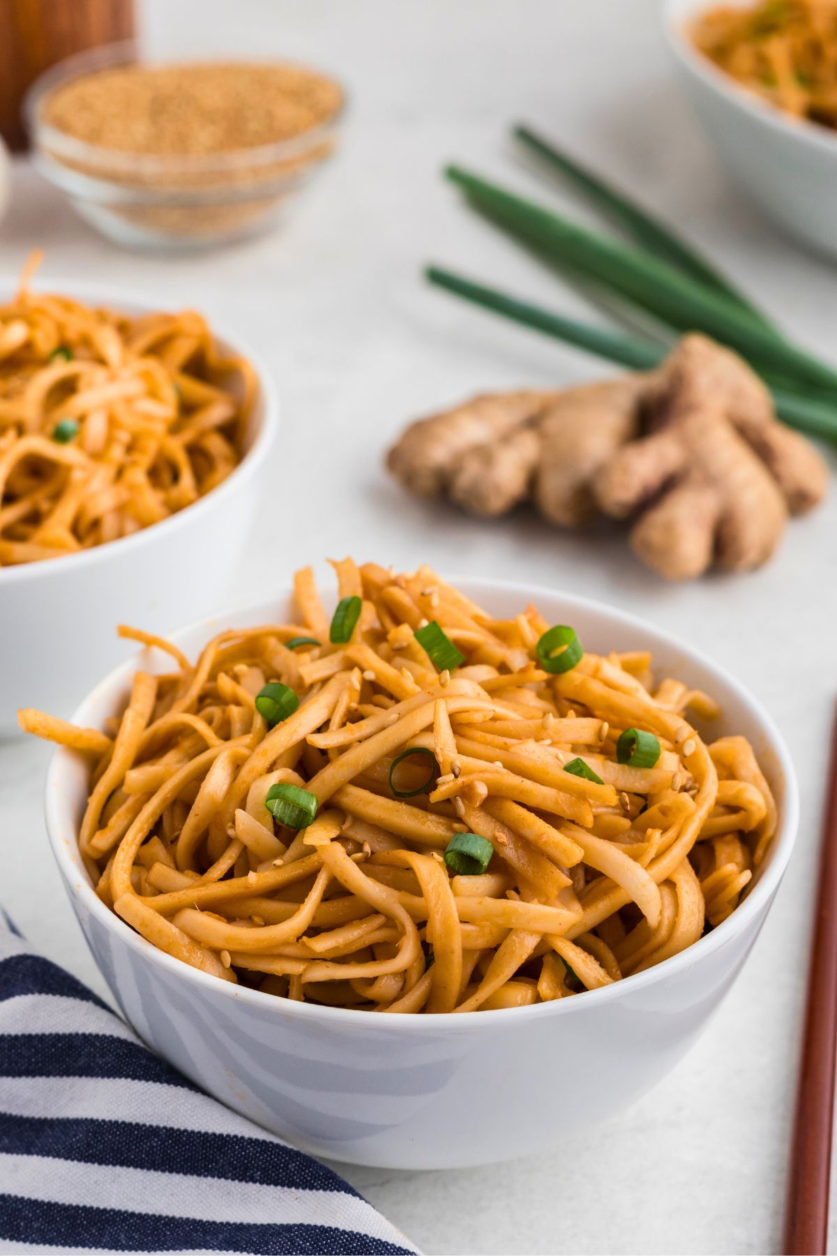 Juicy spicy noodles in a white bowl, garnished with green onions next to a blue and white striped linen. 